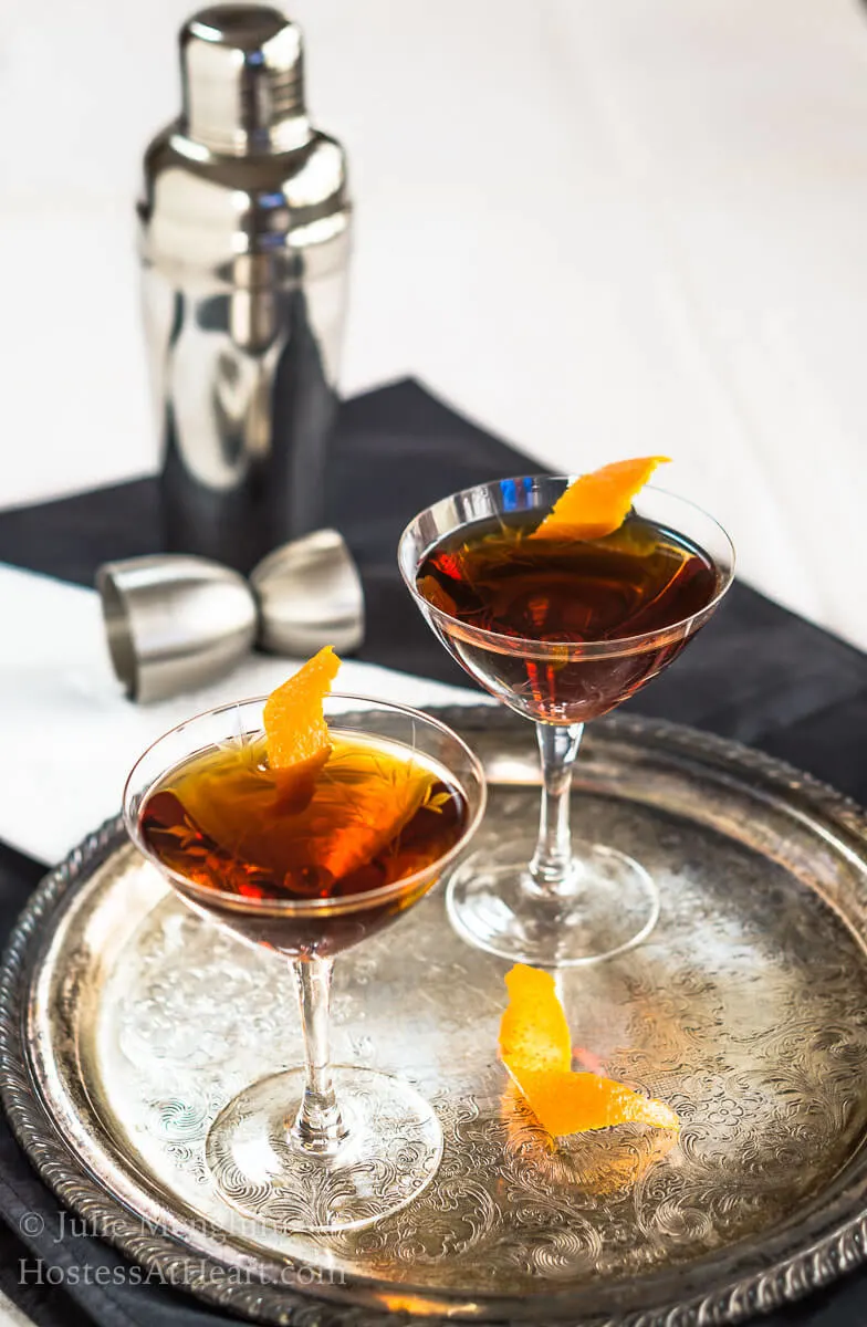 Two glasses of a whiskey cocktail sitting on a silver platter garnished with orange peels. The shaker and shot glass sit in the back.