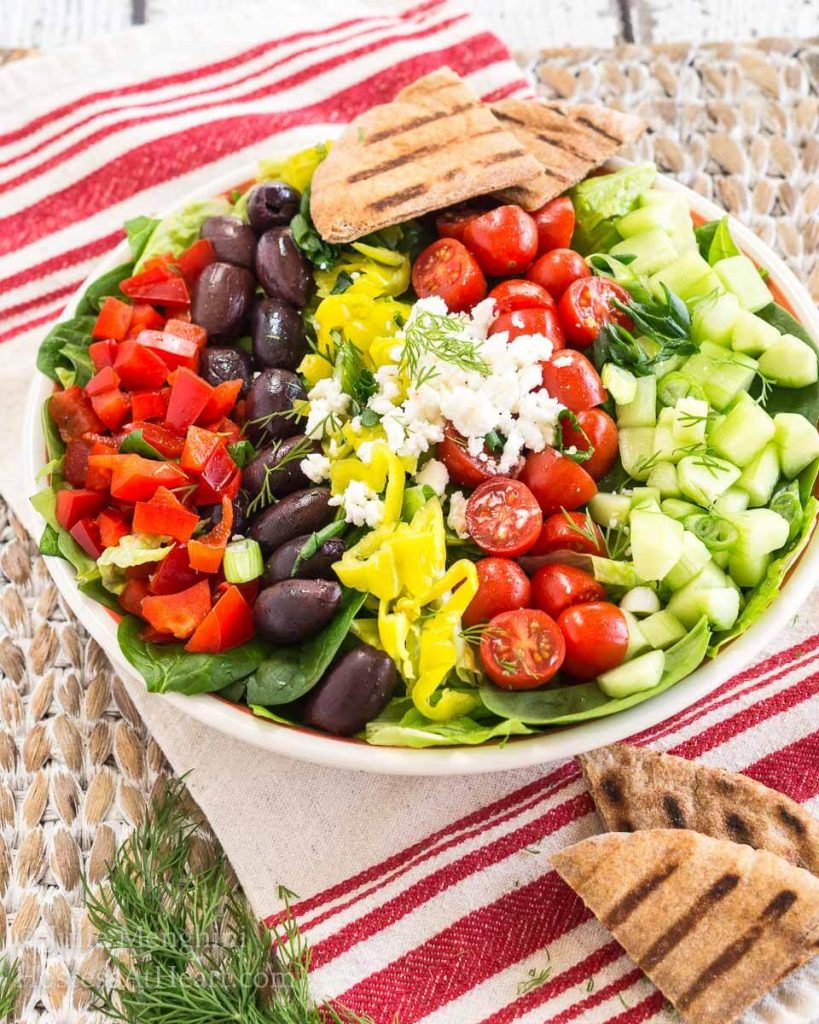 A large bowl of salad topped with diced cucumbers, tomatoes, kalamata olives, red peppers, pepperoncini, and feta cheese. A couple of pita triangles sit in the background.