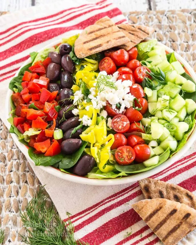 A large bowl of salad topped with diced cucumbers, tomatoes, kalamata olives, red peppers, pepperoncini, and feta cheese. A couple of pita triangles sit in the background.