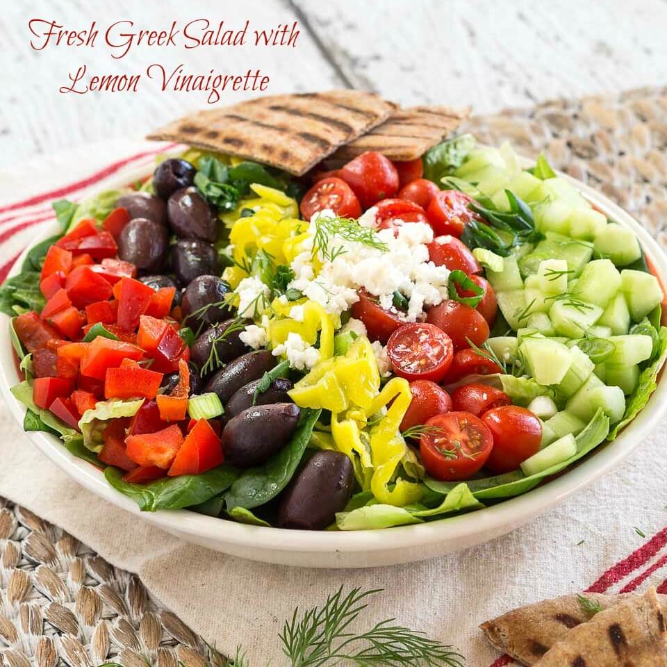 A large bowl of salad topped with diced cucumbers, tomatoes, kalamata olives, red peppers, pepperoncini, and feta cheese. A couple of pita triangles sit in the background.