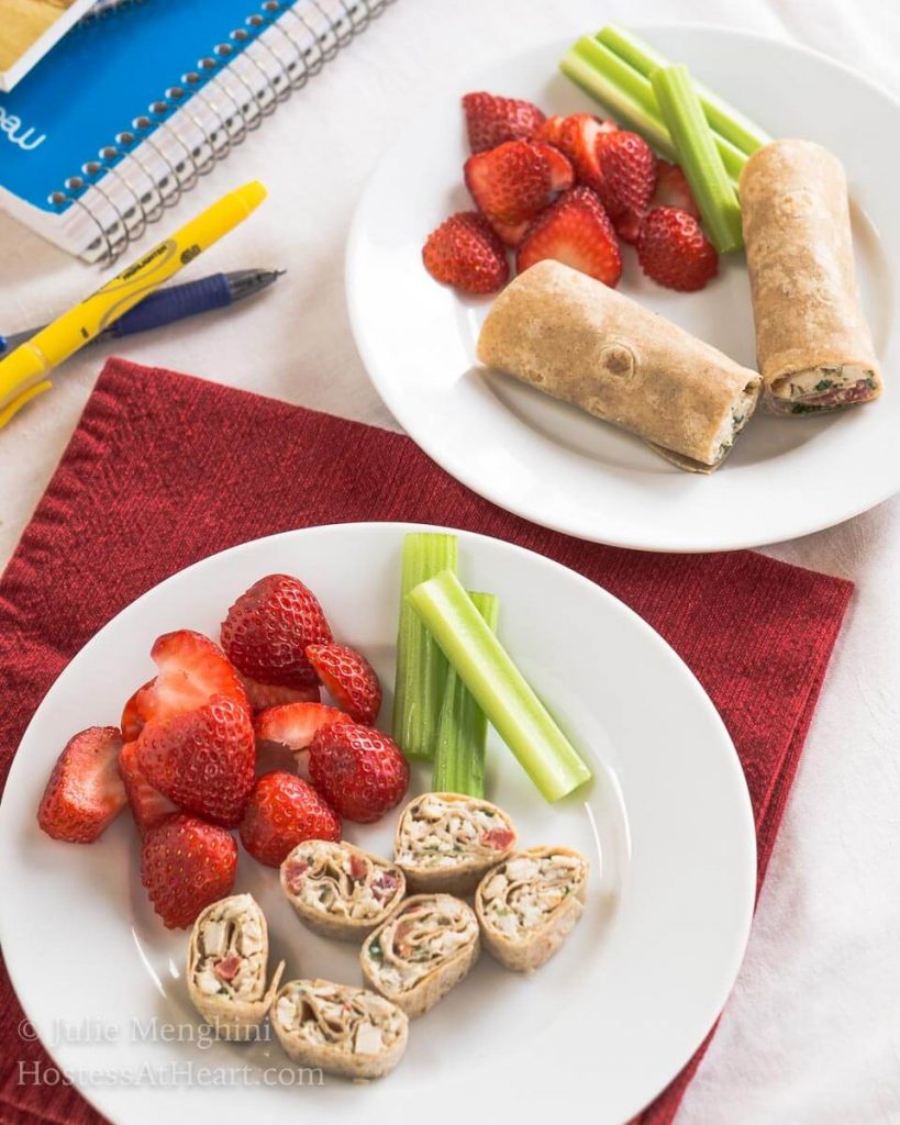 A plate of pinwheels made with sliced chicken and vegetable wrap. Strawberries and stalks of celery sit next to the pinwheels. A plate with the uncut wrap sits in the back.