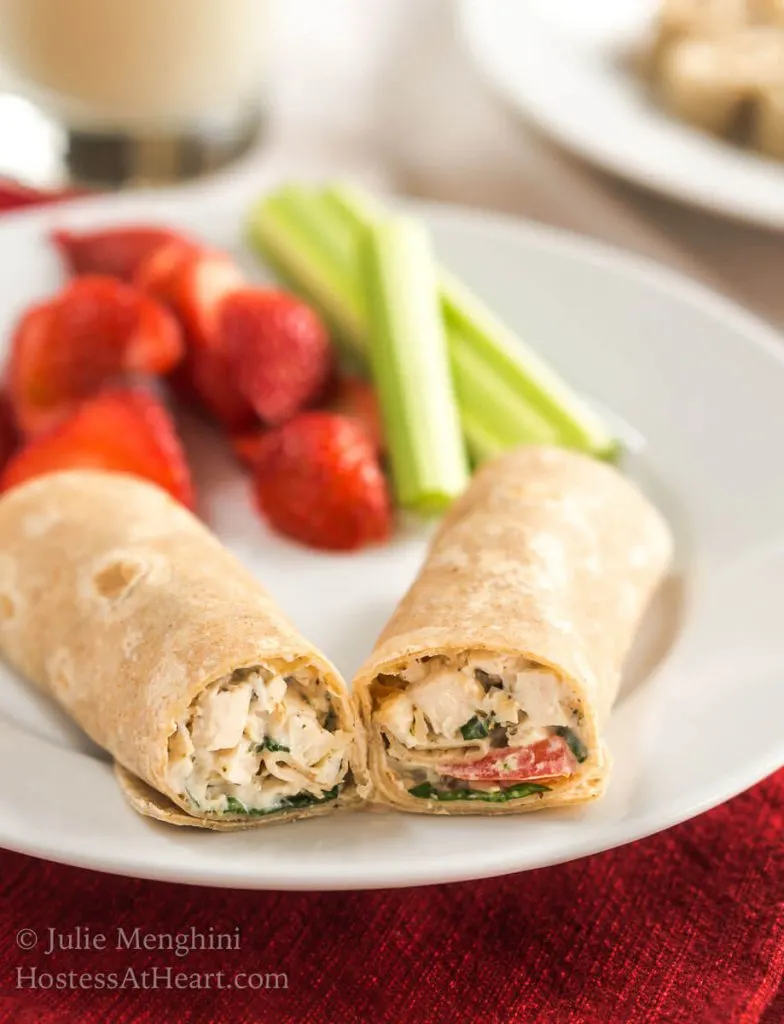 Two chicken wraps on a white plate with sliced strawberries and stalks of celery.