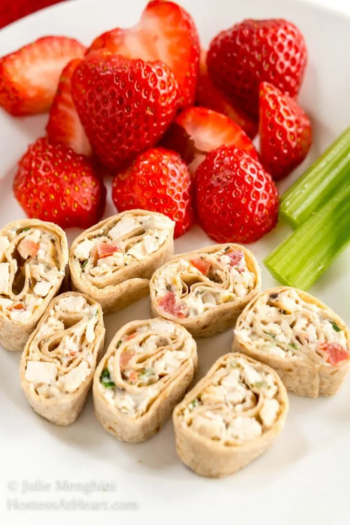A plate of pinwheels made with sliced chicken and vegetable wrap. Strawberries and stalks of celery sit next to the pinwheels.