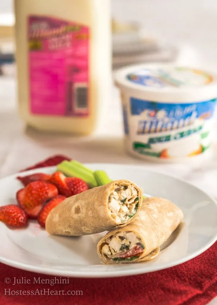 A white plate holding a chicken wrap with slices of strawberries and stalks of celery. A quart of milk and a container of sour cream sits in the background.