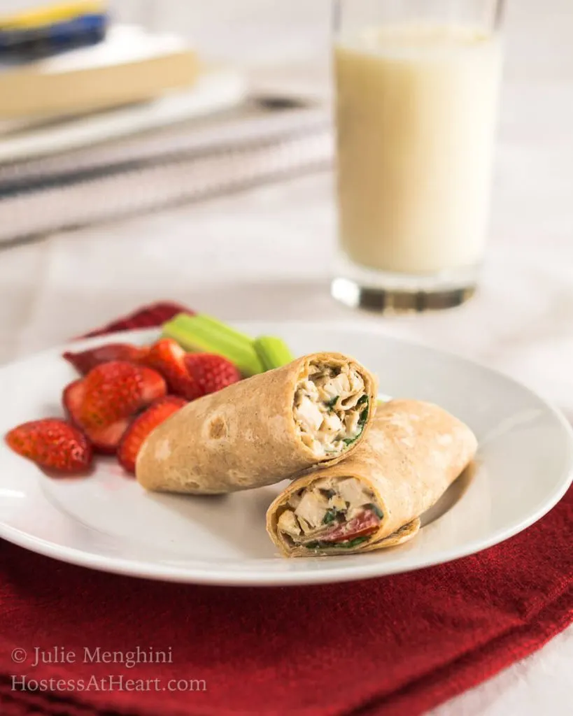 A white plate holding a chicken wrap with slices of strawberries and stalks of celery. A glass of milk sit in the background.