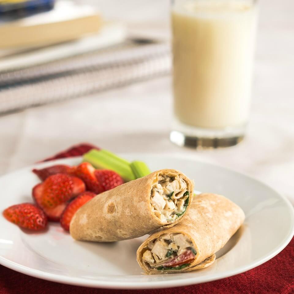 A chicken wrap cut in half on a white plate. A side of strawberries sit in the back of the plate. A glass of milk is in the background.