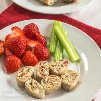 A plate of pinwheels made with sliced chicken and vegetable wrap. Strawberries and stalks of celery sit next to the pinwheels.
