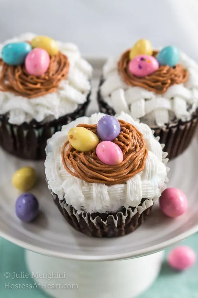Close up of hree cupcakes on a cake stand that are decorated with white basketweave under a piped birdnest and three candy eggs. The stand sits on a soft green napkin and additional candy eggs are sprinkled around the photo.