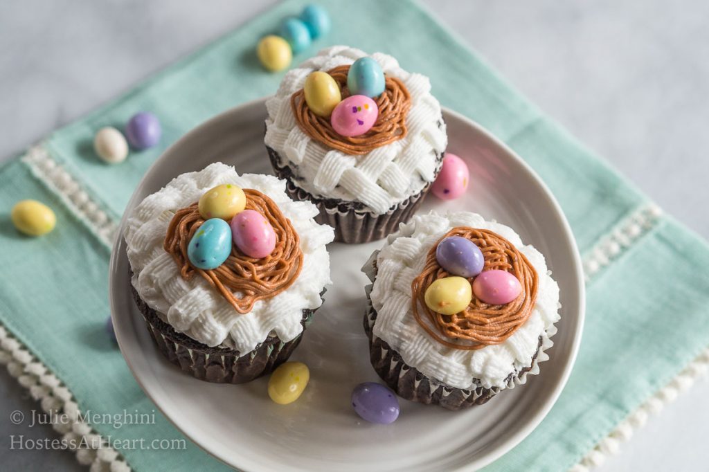 Three cupcakes on a cake stand that are decorated with white basketweave under a piped birdnest and three candy eggs. The stand sits on a soft green napkin and additional candy eggs are sprinkled around the photo.