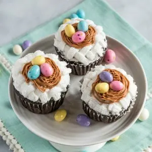 Three cupcakes on a cake stand that are decorated with white basketweave under a piped birdnest and three candy eggs. The stand sits on a soft green napkin and additional candy eggs are sprinkled around the photo.