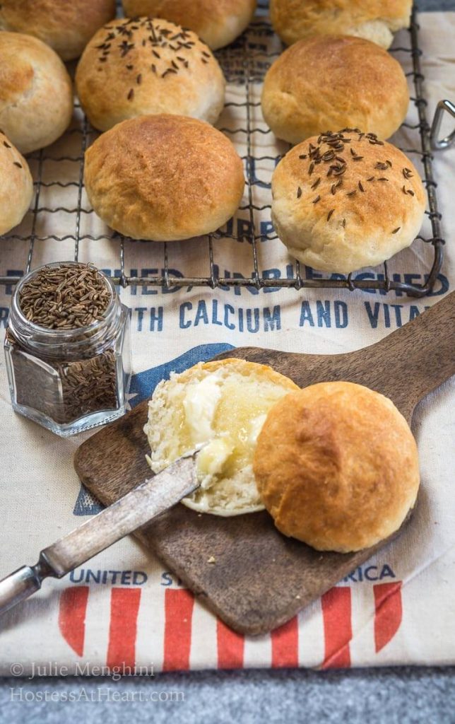 A potato roll sitting on an antique butter paddle that\'s been cut in half and slathered with melted butter. A cooling rack with rolls sits behind the roll, some garnished with caraway seed.