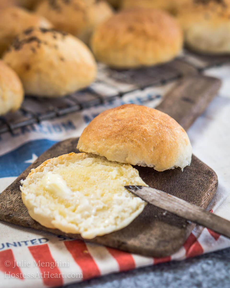 A potato roll sitting on an antique butter paddle that's been cut in half and slathered with melted butter. A cooling rack with rolls sits behind the roll.