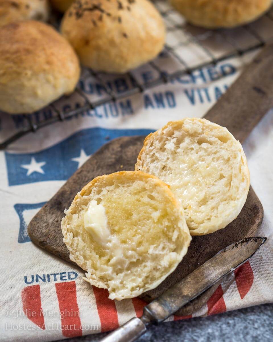 A potato roll sitting on an antique butter paddle that\'s been cut in half and slathered with melted butter. A cooling rack with rolls sits behind the roll.