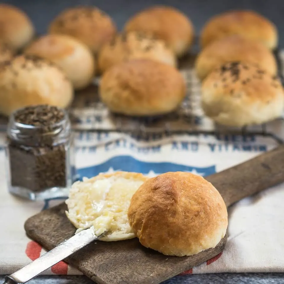 A potato roll sitting on an antique butter paddle that\'s been cut in half and slathered with melted butter. A cooling rack with rolls sits behind the roll.
