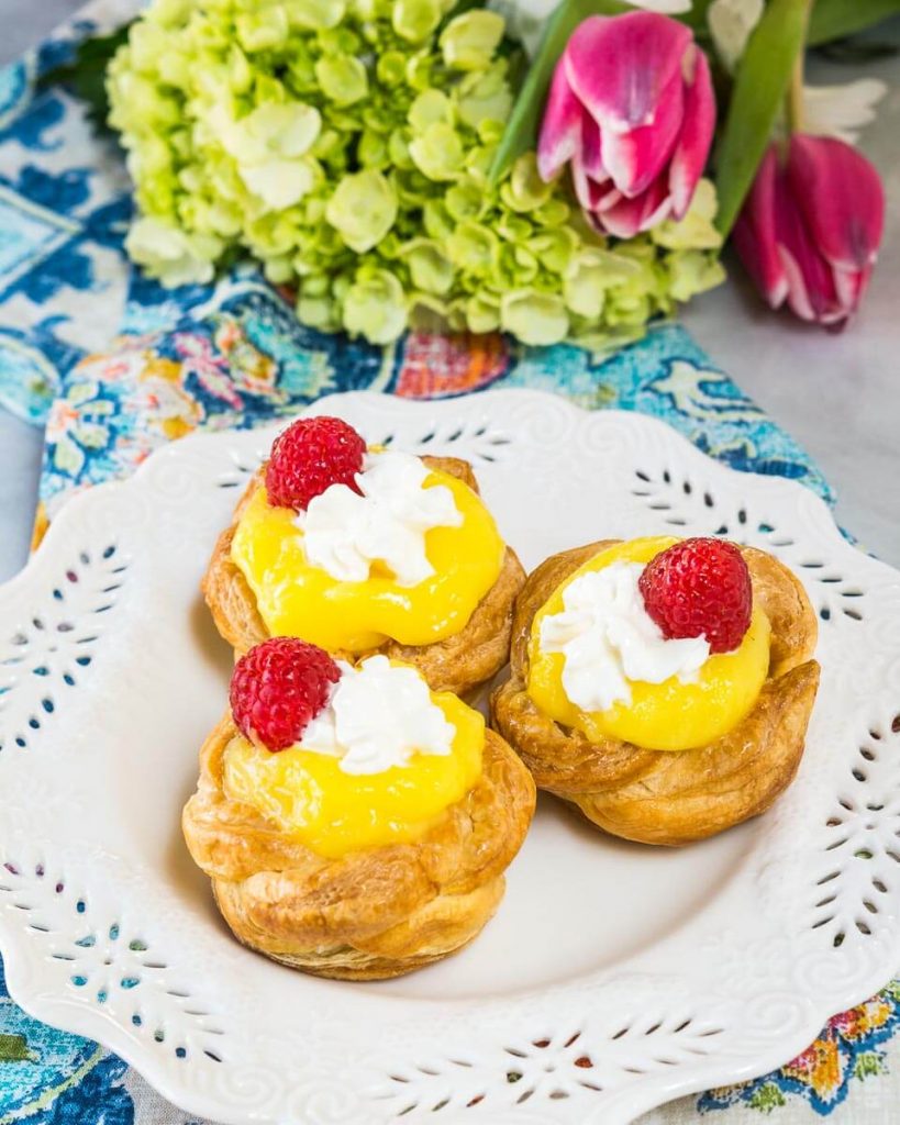 Three Puff Pastry Baskets filled with lemon curd on a white plate. A cup of coffee and a bouquet of flowers sits in the background.