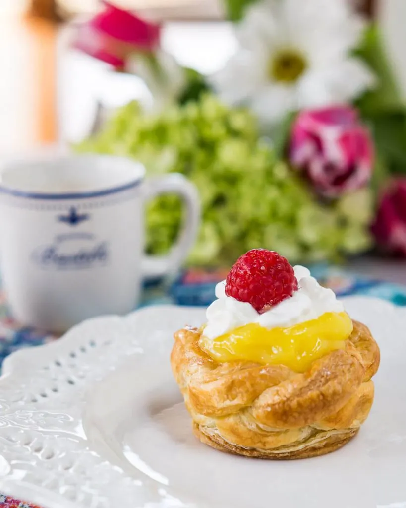 A puff Pastry Basket filled with lemon curd on a white plate. A cup of coffee and a bouquet of flowers sits in the background.