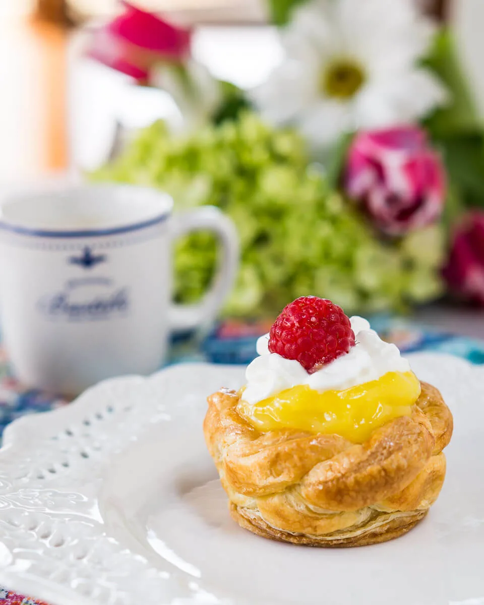 Side view of a puff pastry basket filled with lemon curd topped with whipped topping and a raspberry. A cup of coffee and spring flowers sit in the background.