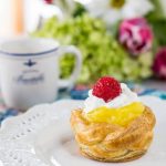 A puff Pastry Basket filled with lemon curd on a white plate. A cup of coffee and a bouquet of flowers sits in the background.