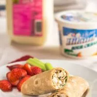 A white plate holding a chicken wrap with slices of strawberries and stalks of celery. A quart of milk and a container of sour cream sits in the background.