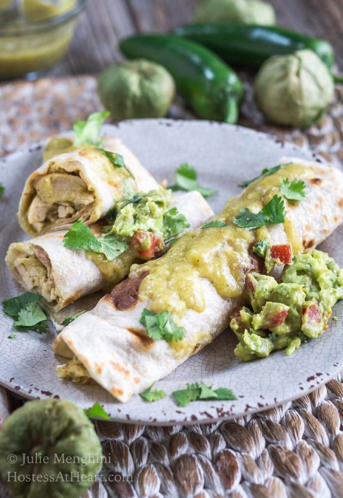 Chicken enchiladas with a green Mole sauce on a gray plate garnished with guacamole and cilantro. Tomatillos and jalapenos sit in the background.