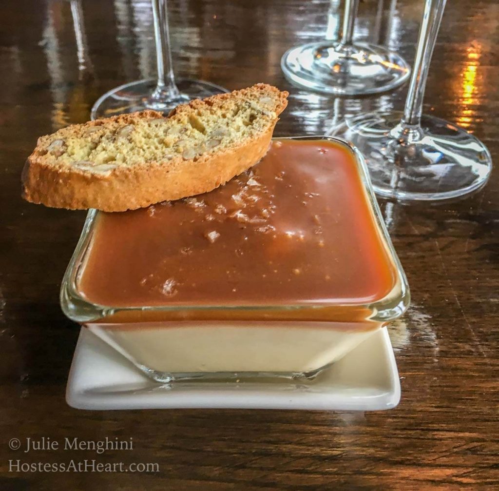 A white bowl of Caramel-topped Budino pudding with a piece of biscotti sitting on the corner of the bowl.