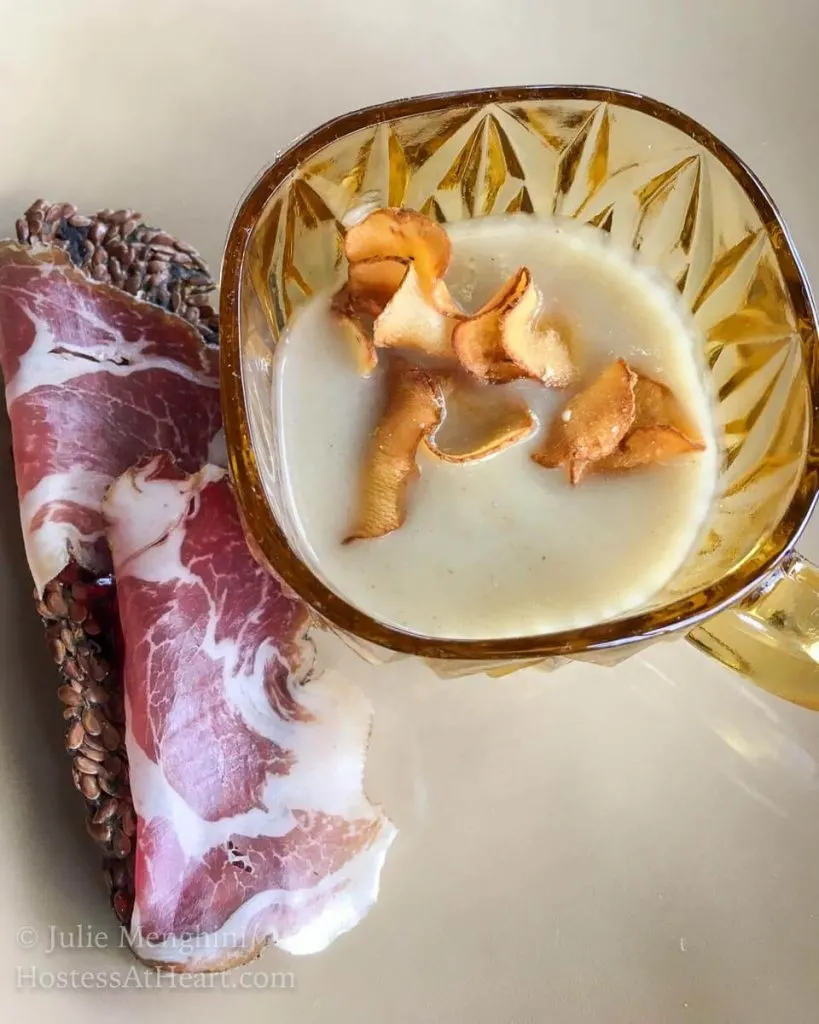Homemade mushroom soup in a gold bowl next to a rustic cracker topped with prosciutto.