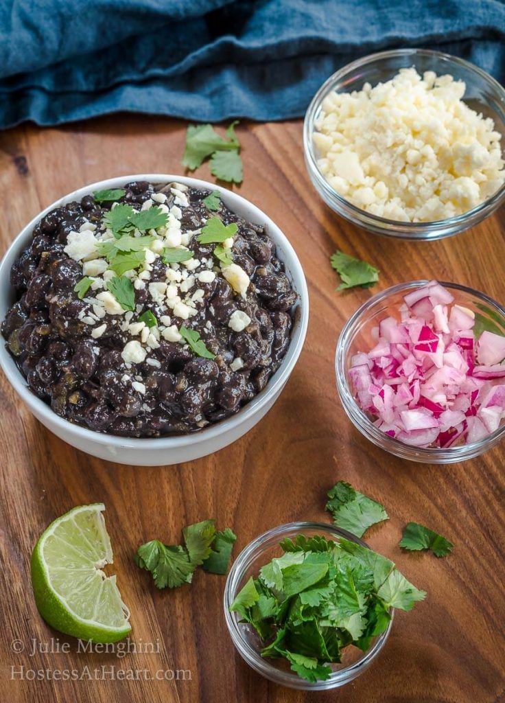 A bowl of black beans garnished with Cotija Cheese and cilantro. Dishes of cheese, red onion, and cilantro sit around the black beans with wedges of lime.