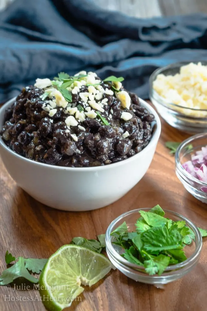 A white bowl filled with black beans garnished with cotija cheese and cilantro. Small dishes of cheese, onions, cilantro, and lime sit around the beans. 