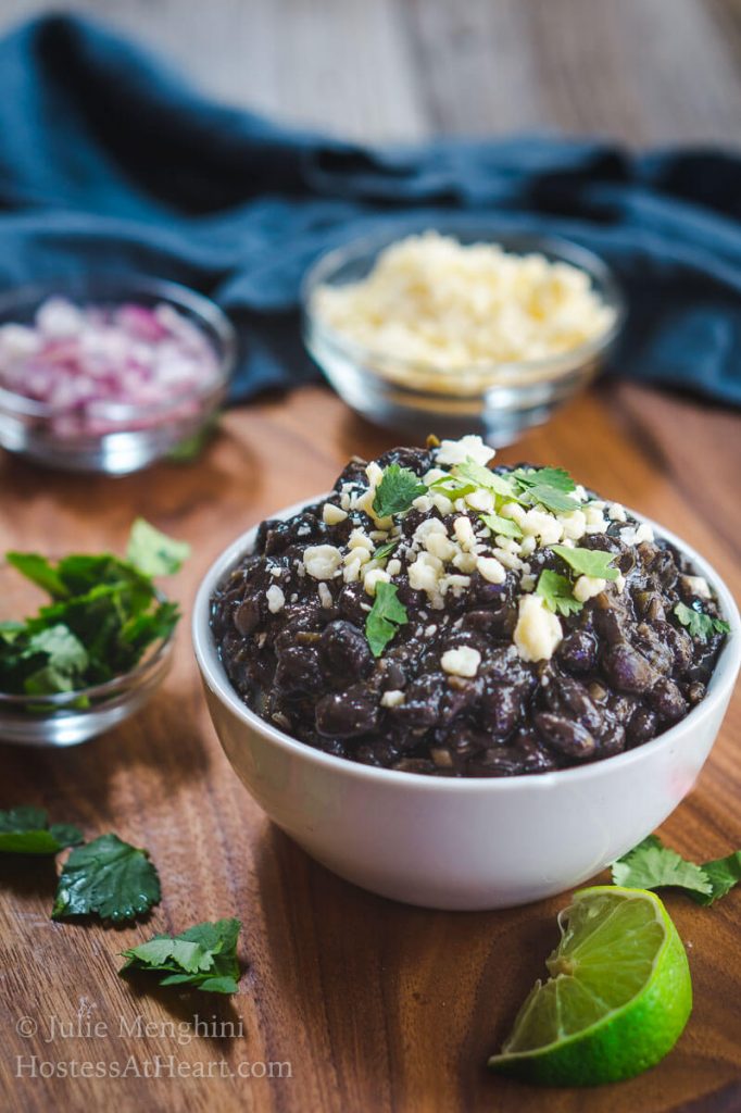 A bowl of black beans garnished with Cotija Cheese and cilantro. Dishes of cheese, red onion, and cilantro sit behind the black beans with wedges of lime.