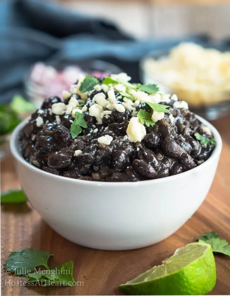 A bowl of black beans garnished with Cotija Cheese and cilantro. Dishes of cheese, red onion, and cilantro sit behind the black beans with wedges of lime.