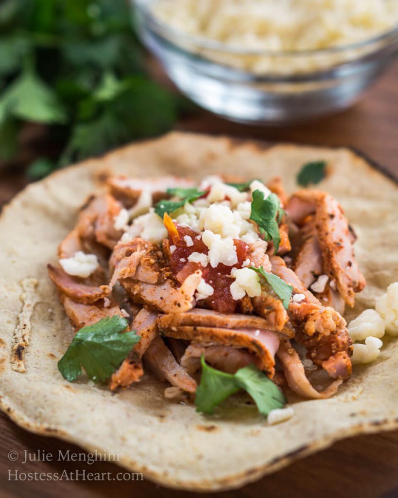 A tortilla filled with Al Pastor pork, Cotija cheese, and cilantro. A dish cheese sits in the background next to fresh cilantro.