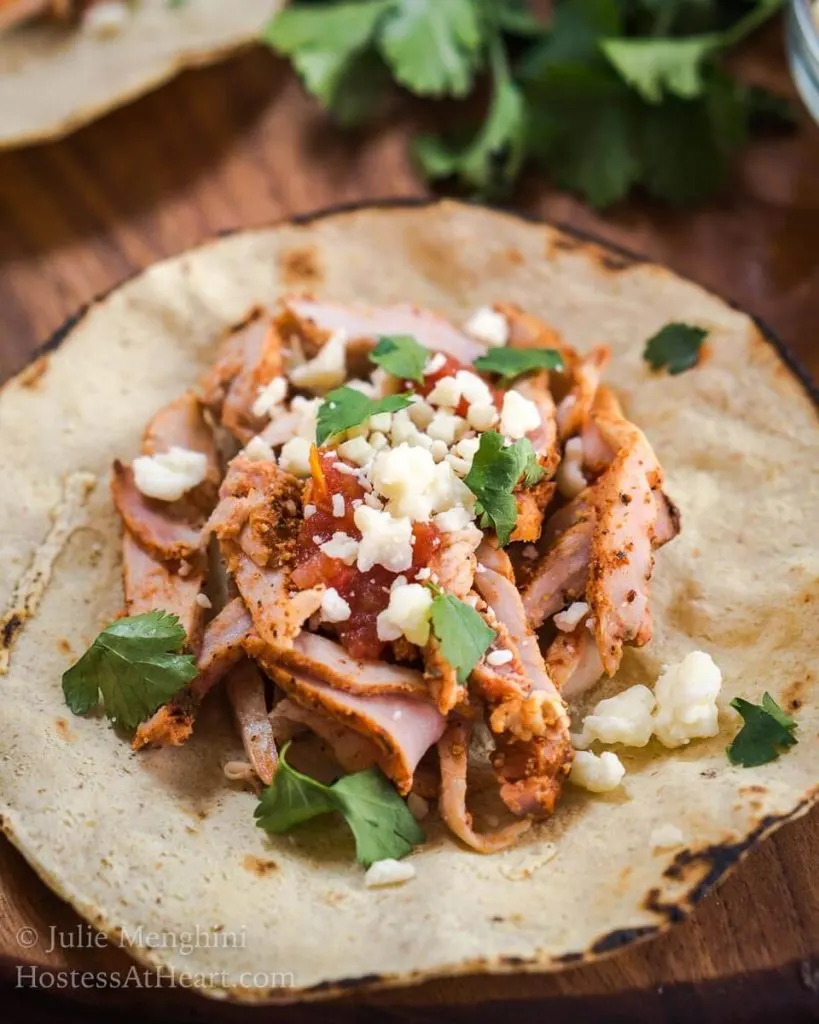 A tortilla filled with Al Pastor pork, Cotija cheese, and cilantro. A dish cheese sits in the background next to fresh cilantro.