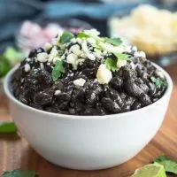 A bowl of black beans garnished with Cotija Cheese and cilantro. Dishes of cheese, red onion, and cilantro sit behind the black beans with wedges of lime.
