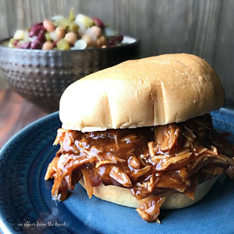 Saucy pulled pork sandwich sitting on a blue plate. A bowl of bean salad sits in the background.