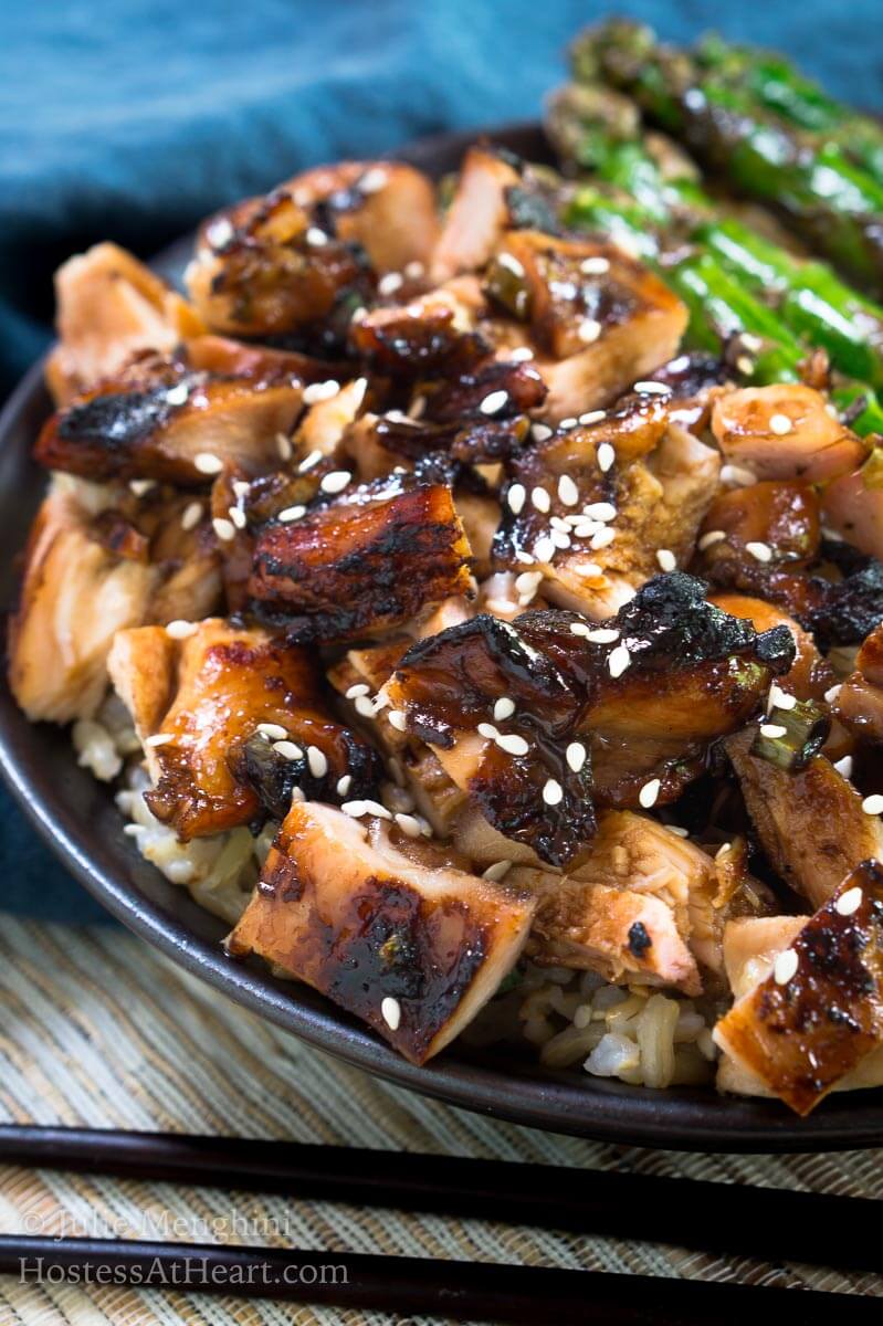 A side view of a gray plate holding diced chicken cooked in teriyaki sauce and sprinkled with sesame seeds sitting in front of grilled asparagus.