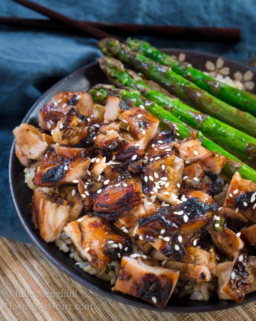 Angle view of a gray plate holding diced chicken cooked in teriyaki sauce in front of grilled asparagus.