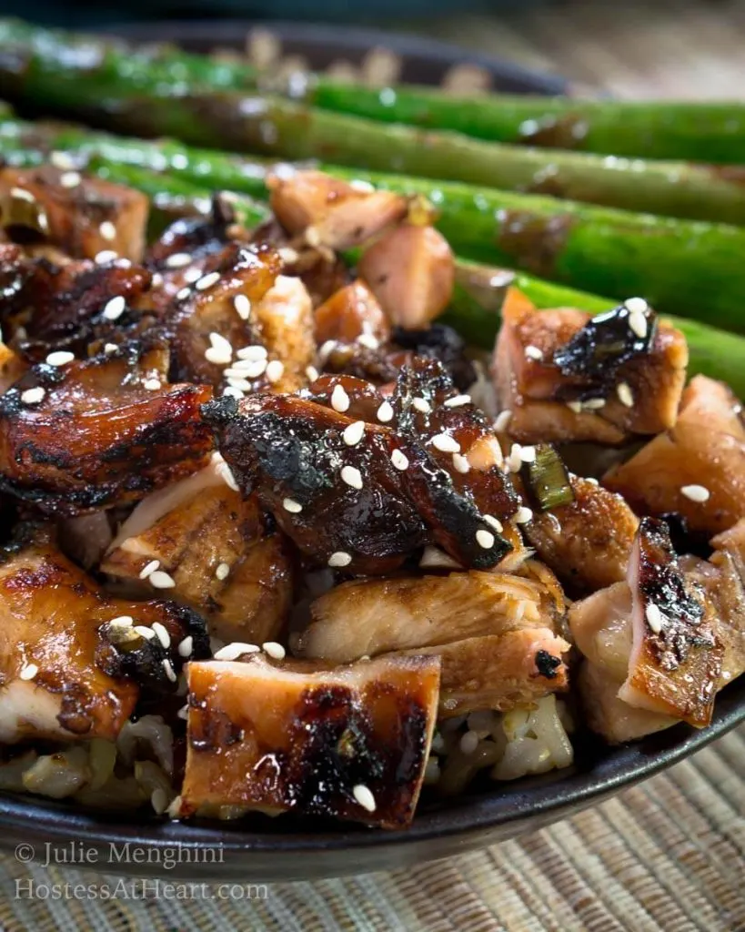 A side view of a gray plate holding diced chicken cooked in teriyaki sauce and sprinkled with sesame seeds sitting in front of grilled asparagus.