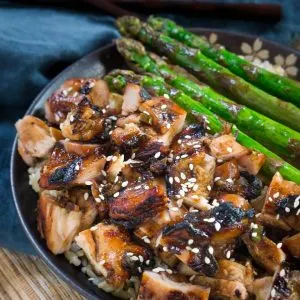 Top view of a bowl of diced chicken marinated and baked in a Teriyaki sauce over rice. Grilled asparagus sit next to the chicken. The bowl sits over a blue napkin.