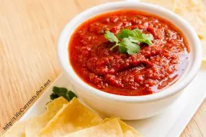 A bowl of Salsa sitting in a white bowl garnished with cilantro. Tortilla chips sit next to the bowl.