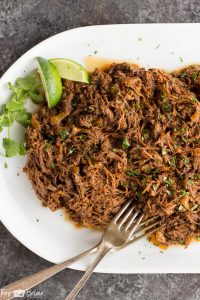 Shredded pork on a white plate with two wedges of lime and cilantro on the side.