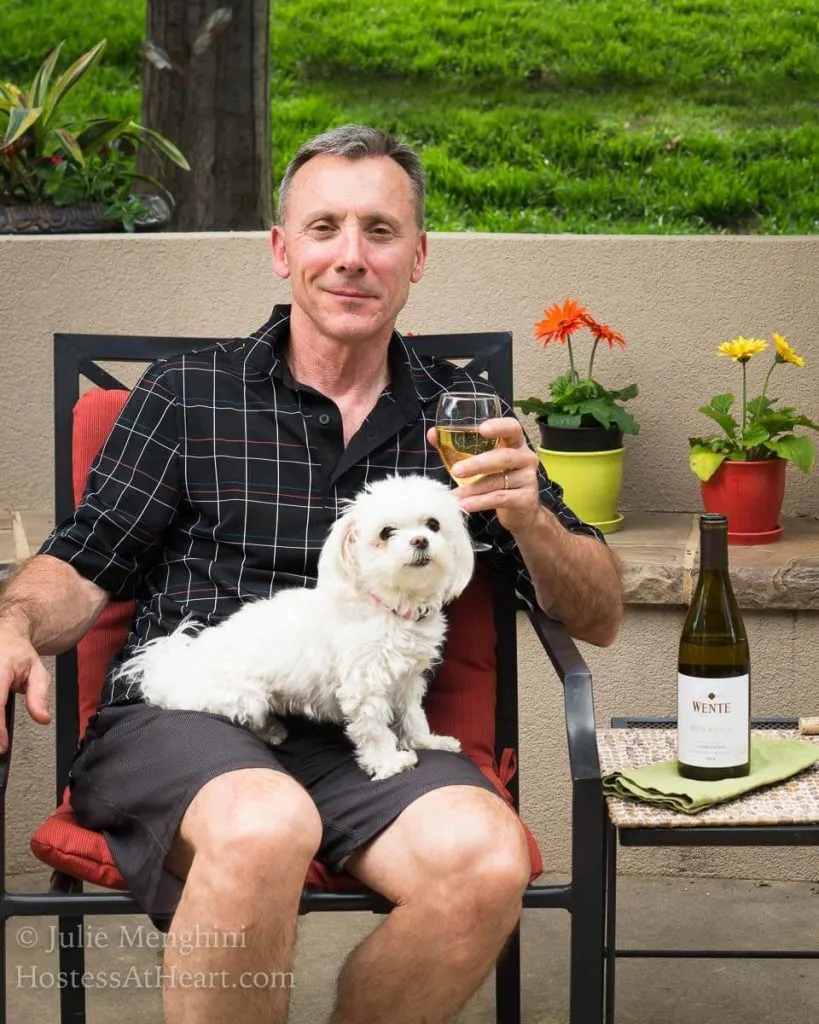 A man holding a glass of Chardonnay with the bottle sitting next to him on a table. A small white dog sits on his lap.