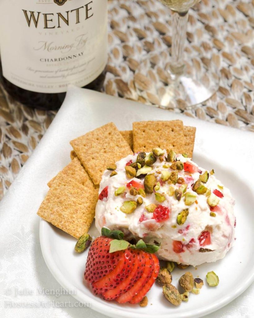 A close up of a white plate holding a cheese ball made with goat cheese, strawberries, and pistachios sitting on a white plate. A bottle of wine sits in the back next to a wine glass.