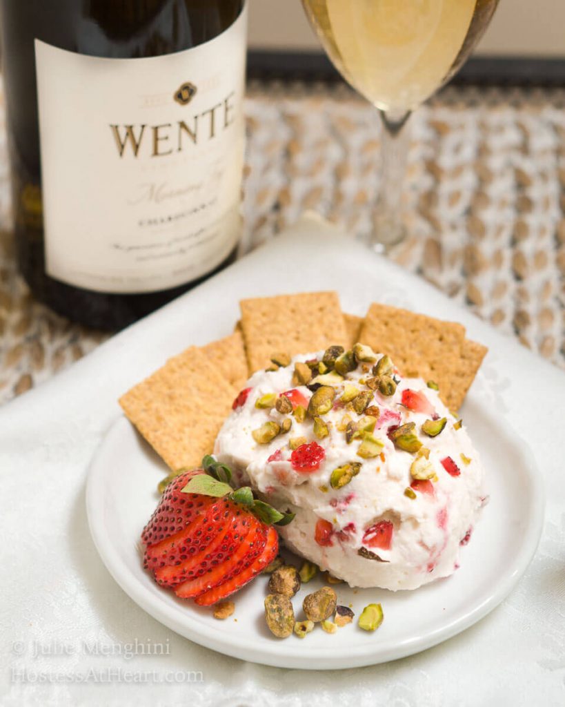 A goat cheese ball made with strawberries and pistachios sitting on a white plate with crackers on the side. A bottle of wine sits in the back.