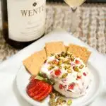 A goat cheese ball made with strawberries and pistachios sitting on a white plate with crackers on the side. A bottle of wine sits in the back.