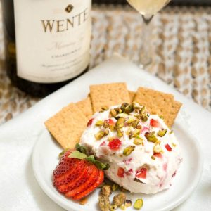 A goat cheese ball made with strawberries and pistachios sitting on a white plate with crackers on the side. A bottle of wine sits in the back.