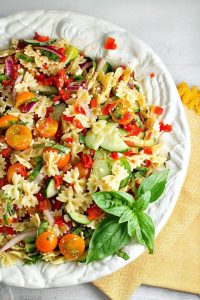 A bowl of bow tie pasta salad filled with tomatoes, red peppers, and basil. A fresh sprig of sweet basil sits to the side.