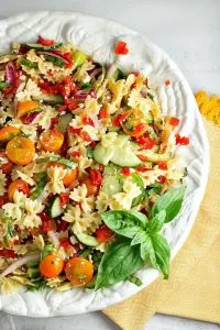 A bowl of bow tie pasta salad filled with tomatoes, red peppers, and basil. A fresh sprig of sweet basil sits to the side.