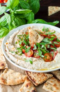 A plate of white bean dip garnished with basil and tomatoes. Pita triangles sit around the bowl.