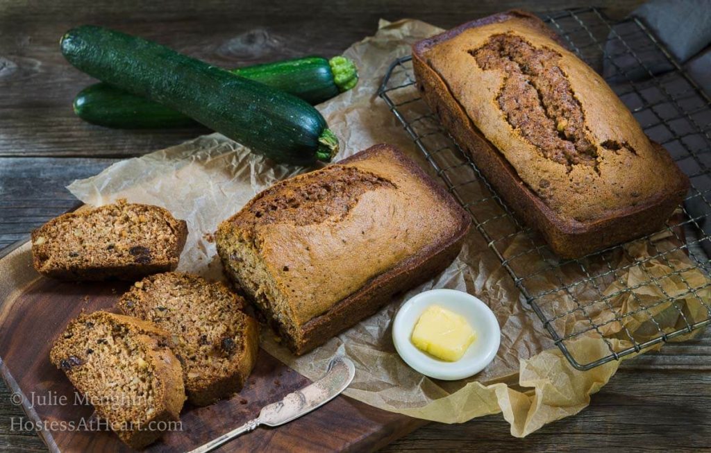 A loaf of zucchini bread sitting on a cooling rack with a sliced loaf sitting next to it. A pad of butter and raw zucchinis sit to the side over parchment paper.