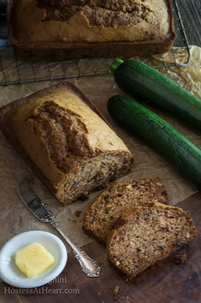 Top angle view of a loaf of sliced zucchini bread. A small white dish holding butter and a knife and two raw zucchini sit to the side.d